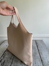 Load image into Gallery viewer, sitting on a table is a bag with a short handle and a short v shaped dip at the center of the bag made in a natural veg tan leather. no patina yet.