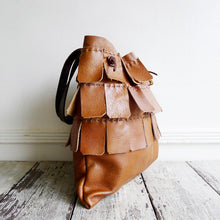 Load image into Gallery viewer, A saffron colored leather bag with three rows of matching wide leather hand sewn fringe covering 1/3 of the small-size bag sits on a table top. Its shoulder length leather straps are a dark brown color.