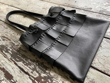 Load image into Gallery viewer, A black leather bag with three rows of wide black leather hand sewn fringe covering 1/3 of the small-size bag starting at the top lays flat on a table top its shoulder.