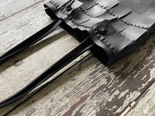 Load image into Gallery viewer, A close up of a black leather bag with three rows of wide black leather hand sewn fringe covering 1/3 of the small-size bag starting at the top lays flat on a table top its shoulder.