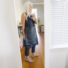 Load image into Gallery viewer, A white haired female stands wearing a narrow and flat rectangular shaped black leather bag over her shoulder. The matching leather strap is tied and knotted to each side of the bag. The bag has a large front pocket. The leather looks supple.