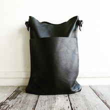 Load image into Gallery viewer, a narrow and flat rectangular shaped black leather bag lays on a table top. The leather looks supple. The black leather strap is tied and knotted to each side of the bag. The front of the bag has a large outside pocket