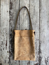 Load image into Gallery viewer, a narrow and flat rectangular shaped tan suede leather bag lays on a table top. The suede looks supple. The dark brown leather strap is tied and knotted to each side of the bag. The front of the bag has a large outside pocket.