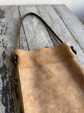 Load image into Gallery viewer, a narrow and flat rectangular shaped tan suede leather bag lays on a table top. The suede looks supple. The dark brown leather strap is tied and knotted to each side of the bag. The front of the bag has a large outside pocket.