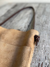 Load image into Gallery viewer, A close up view of the top end of a narrow and flat rectangular shaped tan suede leather bag lays on a table top. The suede looks supple. The dark brown leather strap is tied and knotted to each side of the bag. The front of the bag has a large outside pocket.