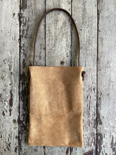 Load image into Gallery viewer, a narrow and flat rectangular shaped tan suede leather bag lays on a table top. The suede looks supple. The dark brown leather strap is tied and knotted to each side of the bag. The front of the bag has a large outside pocket.