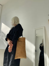 Load image into Gallery viewer, Woman dressed in a long black dress stands wearing a narrow and flat rectangular shape tan suede bag with dark brown straps over her shoulder. The bag has a large front pocket. The suede looks supple.