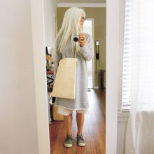 Load image into Gallery viewer, A woman with white hair is standing wearing a narrow and flat rectangular shaped natural veg tan leather bag over her shoulder. The leather patina is a soft peachy blush color. The matching leather strap is tied and knotted to each side of the bag. The front of the bag has a large outside pocket.