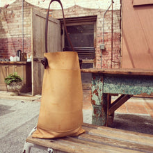 Load image into Gallery viewer, a narrow and flat rectangular shaped natural veg tan leather bag sits on a table top. The leather patina is a light caramel color. The dark brown leather strap is tied and knotted to each side of the bag. The front of the bag has a large outside pocket.