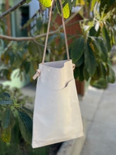 Load image into Gallery viewer, a narrow and flat rectangular shaped natural veg tan leather bag hangs from a tree branch. The leather patina is a soft peachy blush color. The matching leather strap is tied and knotted to each side of the bag. The front of the bag has a large outside pocket.