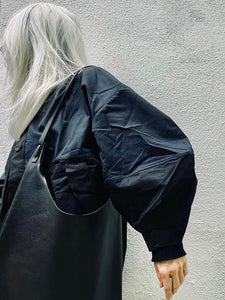Woman is wearing an all-black shoulder length leather bag. The top of the bag dips down into a half-circle shape. Leather straps are tied and knotted at the top left and right sides. The bottom half of the bag is a rectangular shape and the body is flat. The leather looks supple.