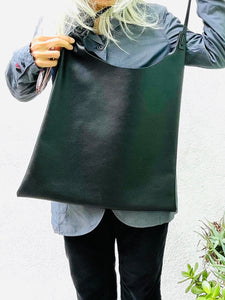 Woman showing an all-black leather bag. The top of the bag dips down into a half-circle shape. Leather straps are tied and knotted at the top left and right sides. The bottom half of the bag is a rectangular shape and the body is flat. The leather looks supple.