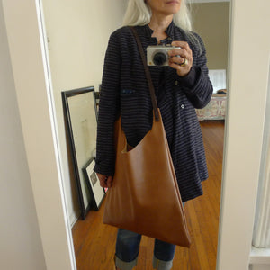 Woman is wearing a crossbody length leather bag. The top of the bag dips down into a half-circle shape. Leather straps are tied and knotted at the top left and right sides. The bottom half of the bag is a rectangular shape and the body is flat. The leather looks supple. This bag can be ordered in black, distressed brown and natural veg tan leather and is no longer available in the pictured tan leather. 