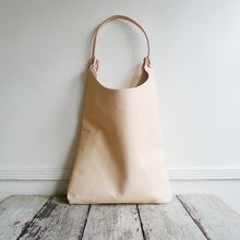 Load image into Gallery viewer, Front view of a shoulder length bag made in natural vegetable tanned leather. The top of the bag dips down into a half-circle shape. Leather straps are tied and knotted at the top left and right sides. The bottom half of the bag is a rectangular shape and the body is flat.
